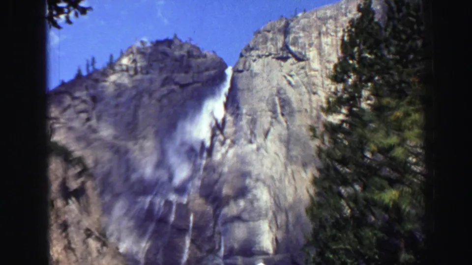 1969 Lake Tahoe 8mm view of a waterfall cascading over a granite wall, with conifers at the right edge and blue sky above.