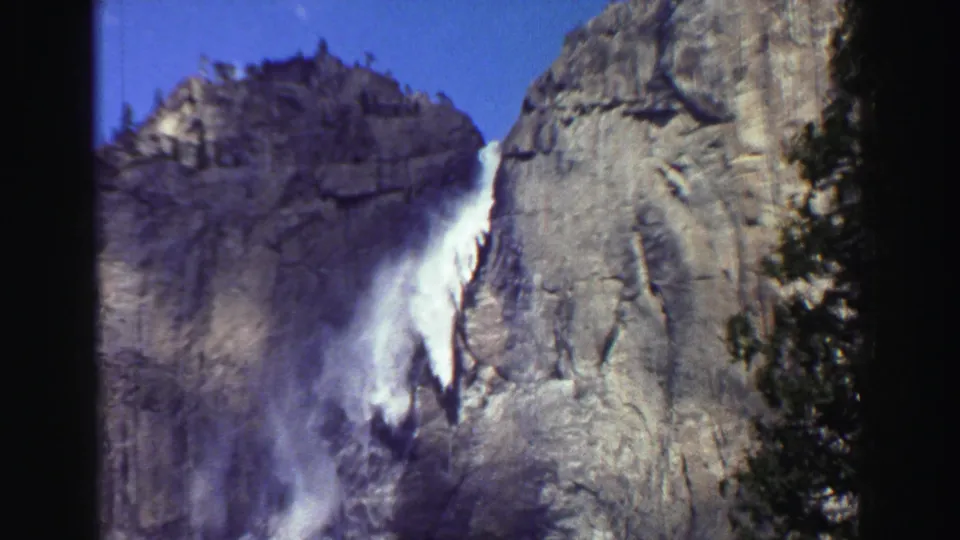 1969 Lake Tahoe home-movie frame with a closer waterfall view against pale granite, bordered by evergreens and visible film softness.