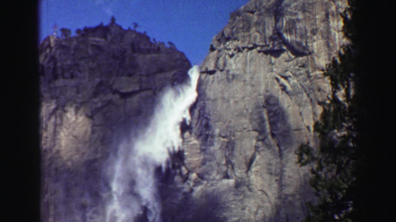 Waterfall dropping beside a granite cliff with dark tree trunks in the foreground.