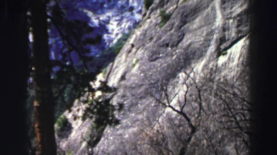 Handheld 1969 8mm view of a Lake Tahoe granite cliff seen through dark trees and leafless branches.