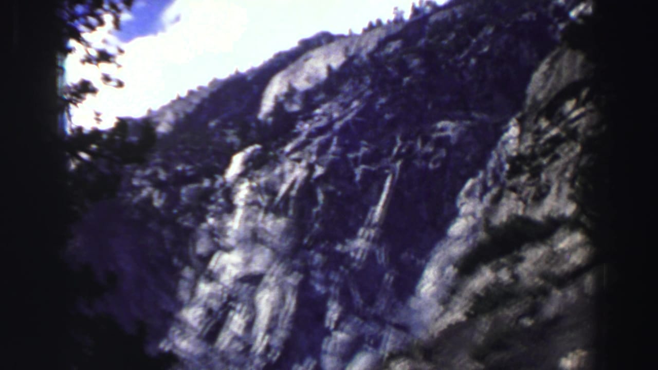Rocky mountain wall and dark tree trunk seen against a bright sky.