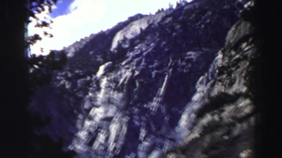 1969 Lake Tahoe home-movie frame with a dark central tree trunk dividing views of rugged granite slopes and sky.