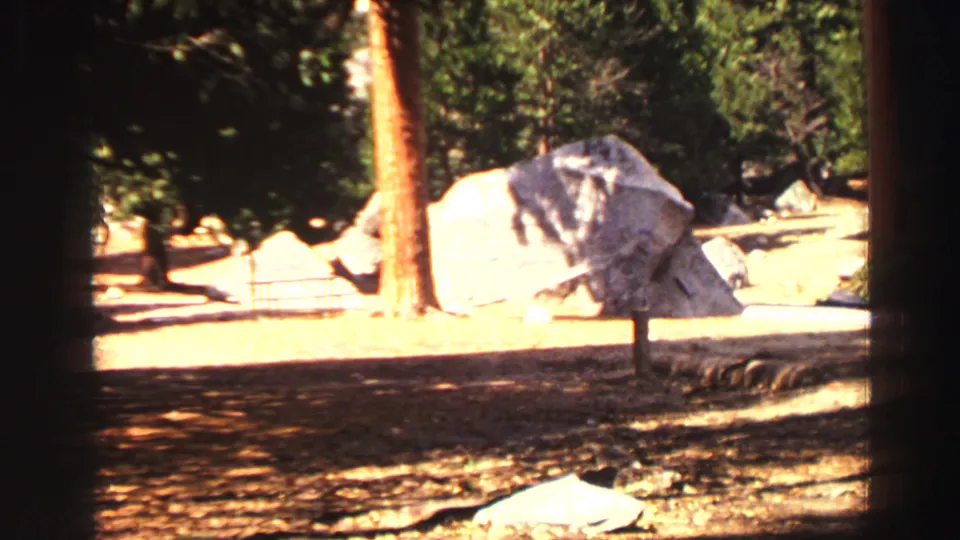 1969 Lake Tahoe park view showing sunlit dirt ground, pine trees, and large granite boulders from a handheld 8mm camera.