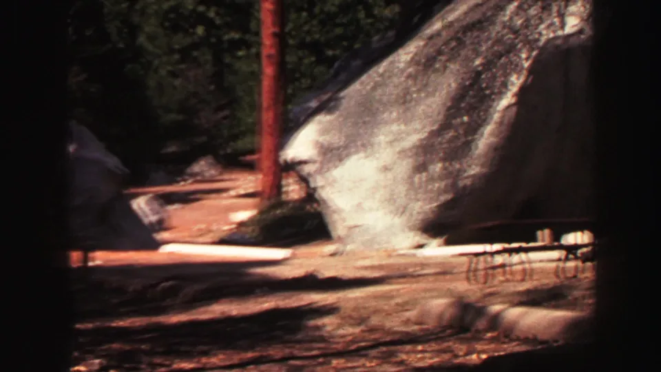 1969 Lake Tahoe 8mm frame with a large granite boulder, pine trunk, shaded ground, and partial picnic furniture at the edge.