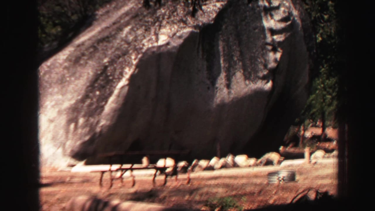 1969 Lake Tahoe 8mm frame with picnic tables, pine trunks, and a broad granite boulder in a forest recreation area.
