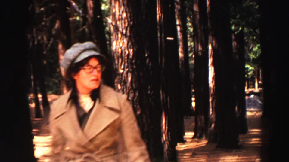 1969 Lake Tahoe 8mm frame showing an adult woman in a cap, glasses, and coat walking through a pine forest.
