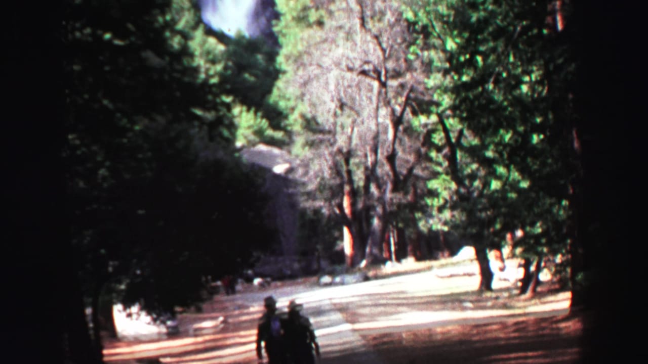 Two adults appear near the lower edge of a wooded path while a waterfall and rocky hillside are partly visible above them.