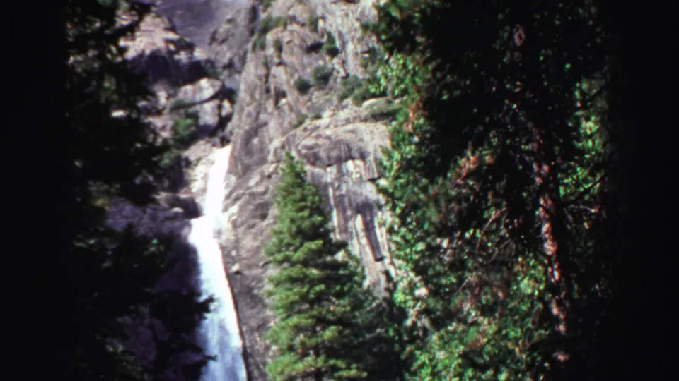 1969 Lake Tahoe home-movie frame showing a bright waterfall on a rocky cliff, bordered by dark evergreens and 8mm film edges.