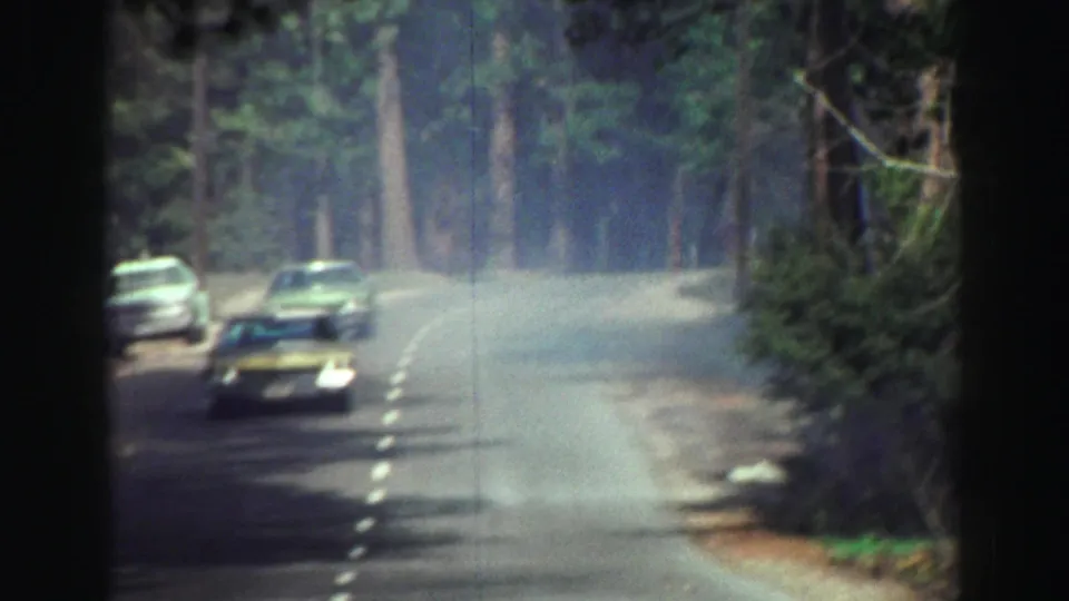 1969 Lake Tahoe frame with two cars on a curving forest road and another car near the roadside shoulder.