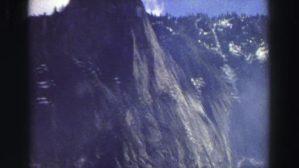 1969 Lake Tahoe home-movie frame showing a steep pale rock face, forested ridge, and scattered snow patches in soft focus.