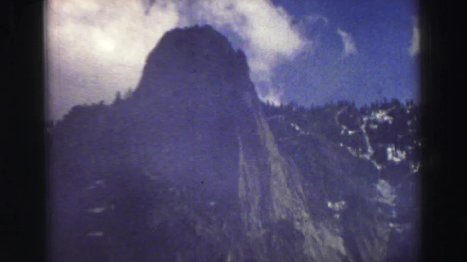 1969 Lake Tahoe frame showing a rounded mountain form, surrounding rocky ridges, snow patches, and a bright clouded sky.