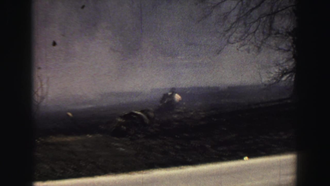 Dark rural ground under windblown haze with bare branches and a distant figure.