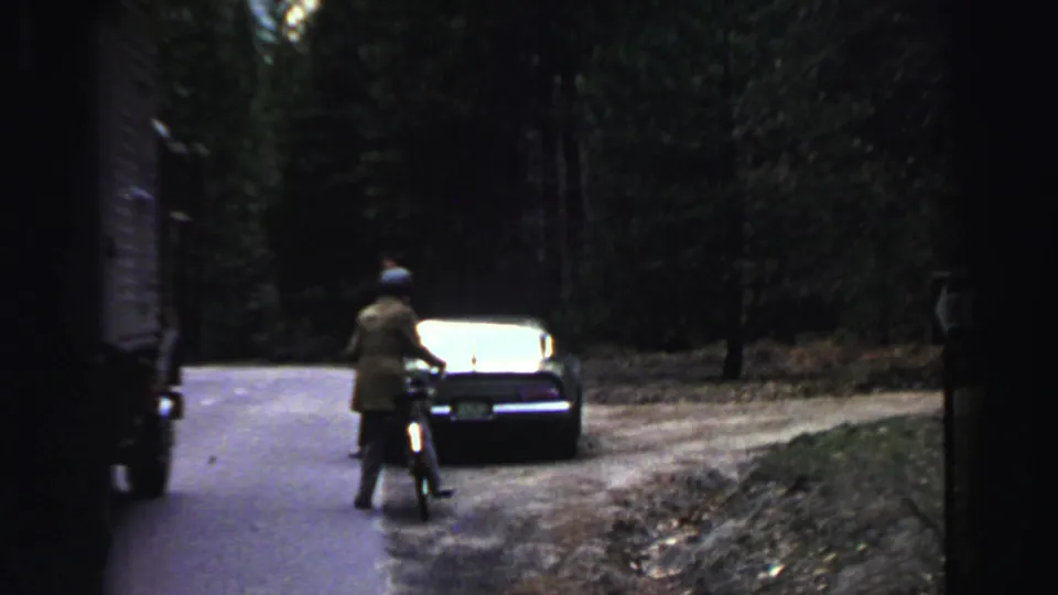 1969 Lake Tahoe home-movie frame showing a cyclist near a dark sedan on a forest road.