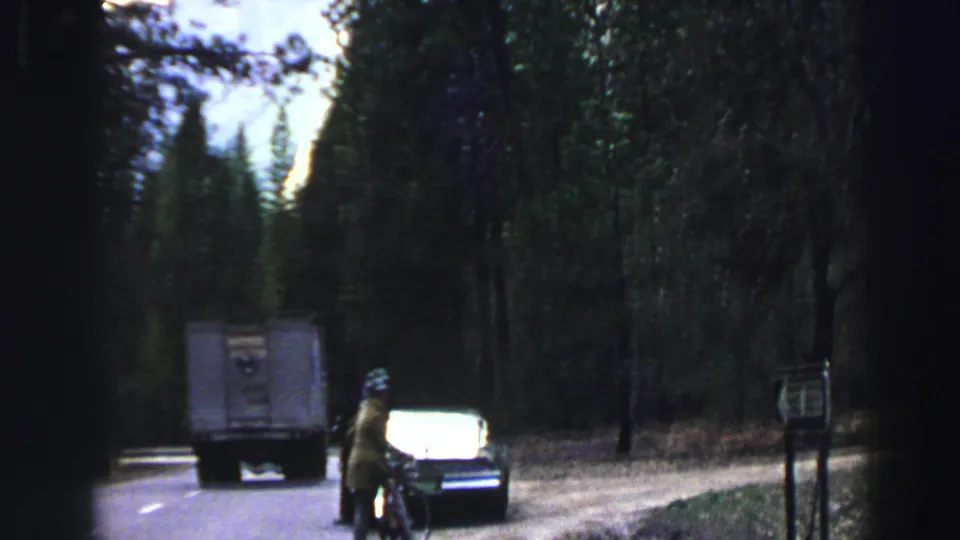 1969 Lake Tahoe 8mm frame of a box truck, cyclist, and dark sedan on a wooded road with roadside signs.