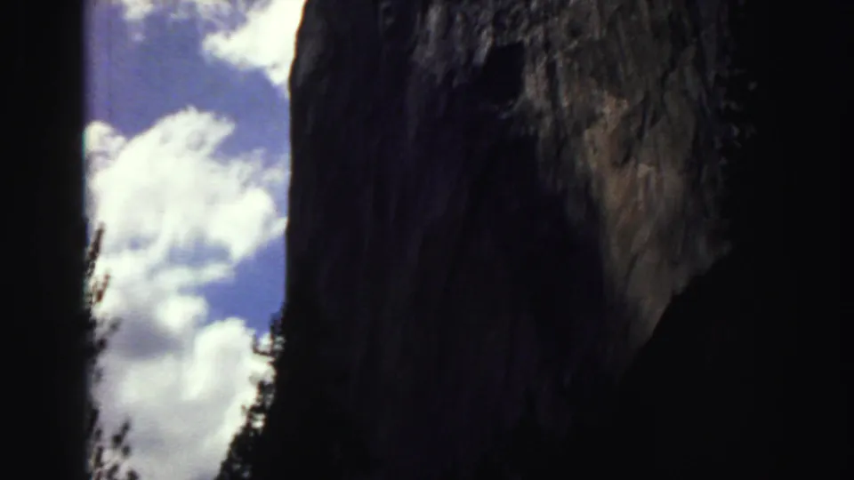 1969 8mm view of a massive granite cliff above shadowed pines, with blue sky and white clouds.