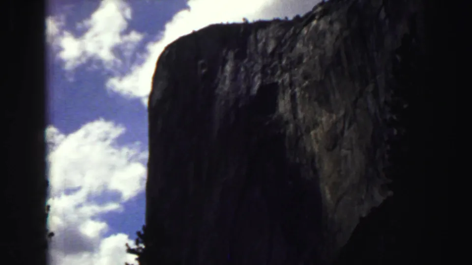 Lake Tahoe home-movie frame showing a sunlit granite cliff edge, dark trees, and bright clouds in blue sky.