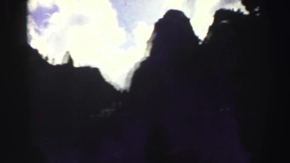1969 Lake Tahoe 8mm frame with shadowed ridgelines, a narrow rock spire, dark tree edges, and glowing clouds.