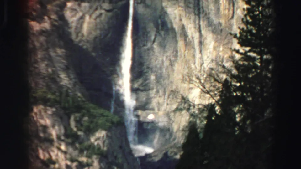 1969 Lake Tahoe travel footage frame showing a vertical waterfall, pale rock wall, evergreen forest, and soft 8mm color grain.