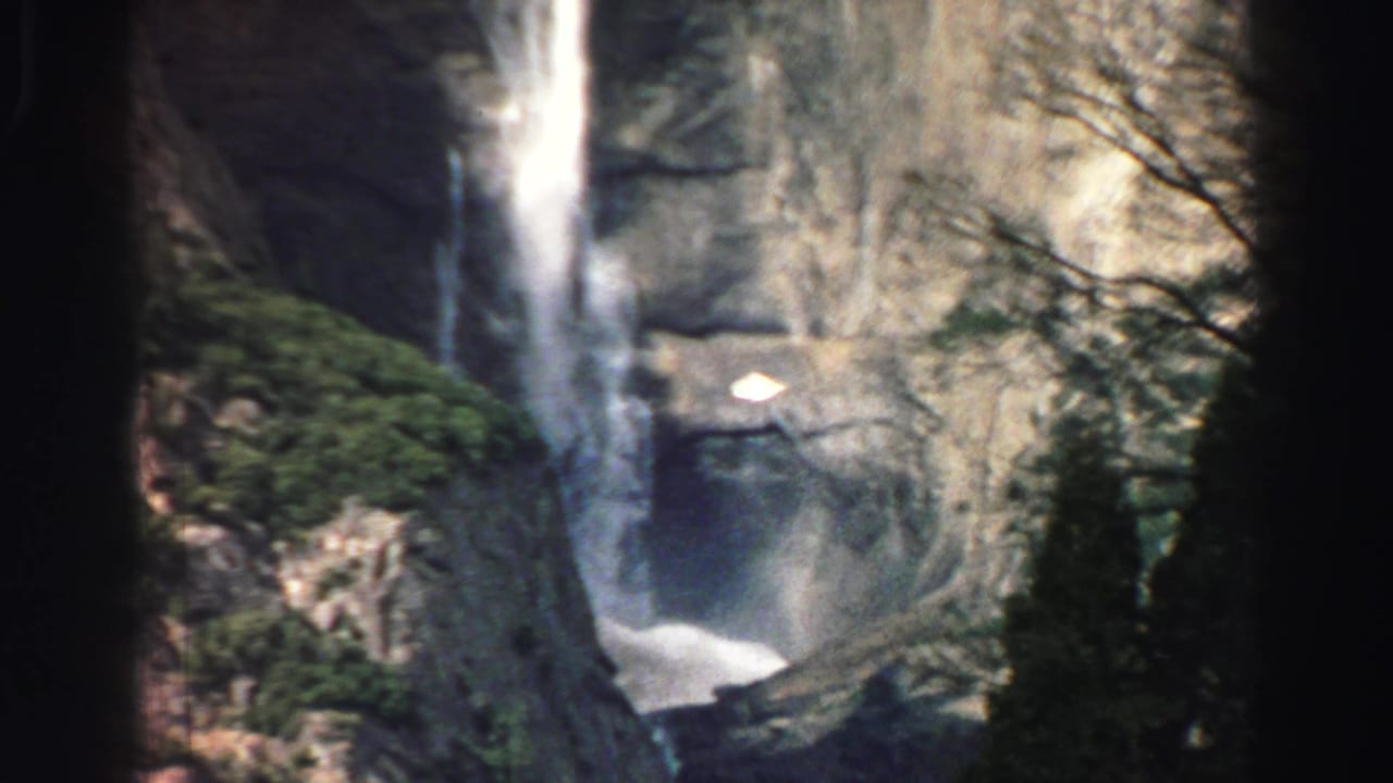Waterfall dropping down a rocky cliff behind dark tree trunks.