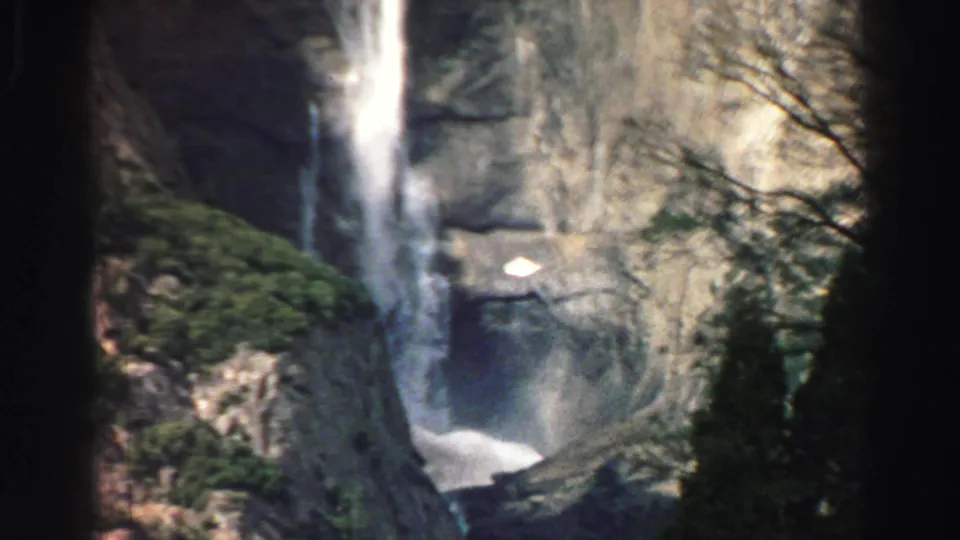1969 Lake Tahoe 8mm frame showing a waterfall descending a rocky cliff, partly obscured by dark foreground tree trunks.