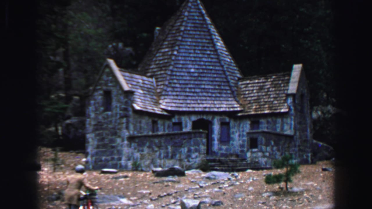Stone building with steep shingled roof in a wooded rocky setting.