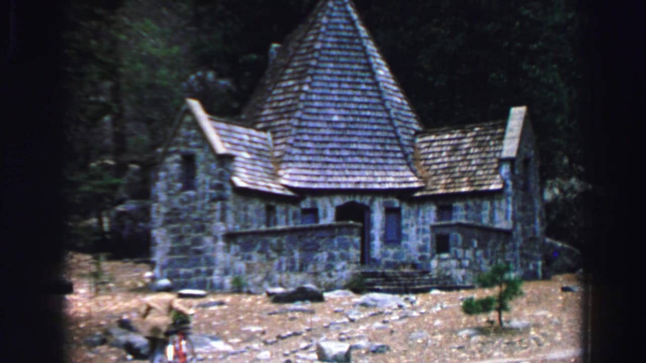 Rustic stone house with steep roof and a person moving near the foreground.