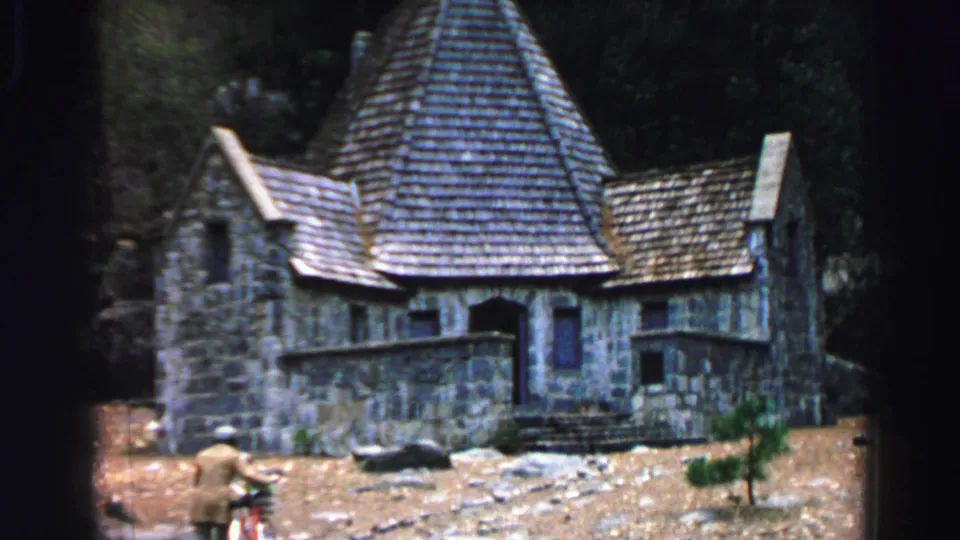 Lake Tahoe 1969 8mm frame showing a person and bicycle closer to the lower left, with a steep-roofed gray stone building and wooded hillside behind.