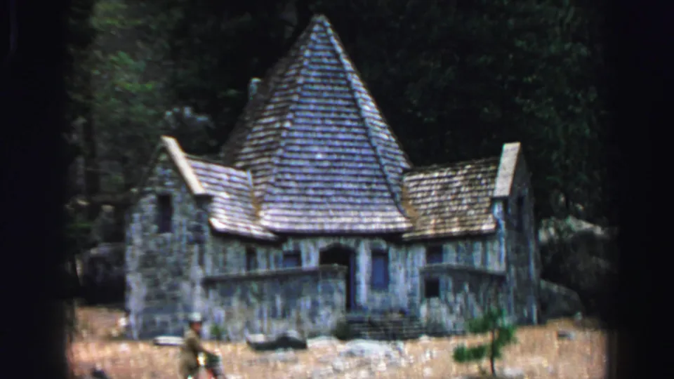 Final 1969 Lake Tahoe frame showing the rustic stone house, steep shingle roof, shaded trees, scattered rocks, and a distant person near a bicycle.