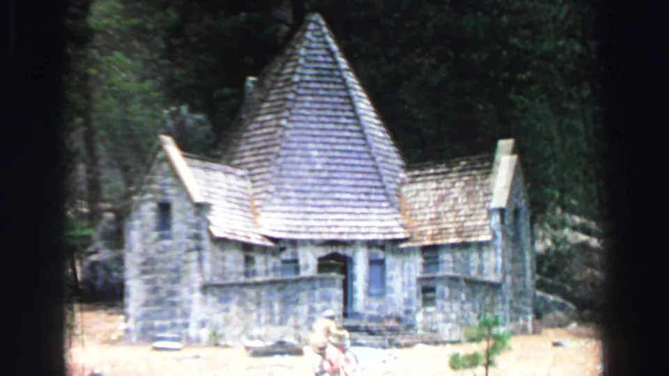 1969 Lake Tahoe 8mm frame showing a rustic stone building with a steep conical roof among trees, with a person near the entrance area.