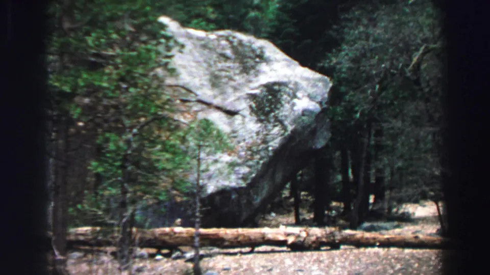 1969 Lake Tahoe 8mm frame focused on a massive angled boulder in dense woods, with a fallen log and brush in front.