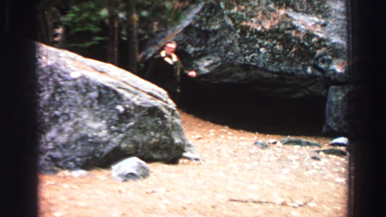 Man in a dark suit standing near large rocks in a wooded area.