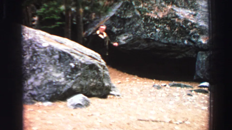 1969 Lake Tahoe home-movie frame showing a suited man beside large boulders on dirt ground under trees.