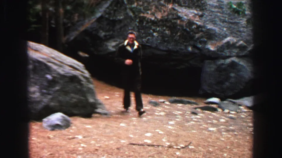 1969 Lake Tahoe frame with a suited man walking across rocky dirt terrain in a wooded outdoor area.