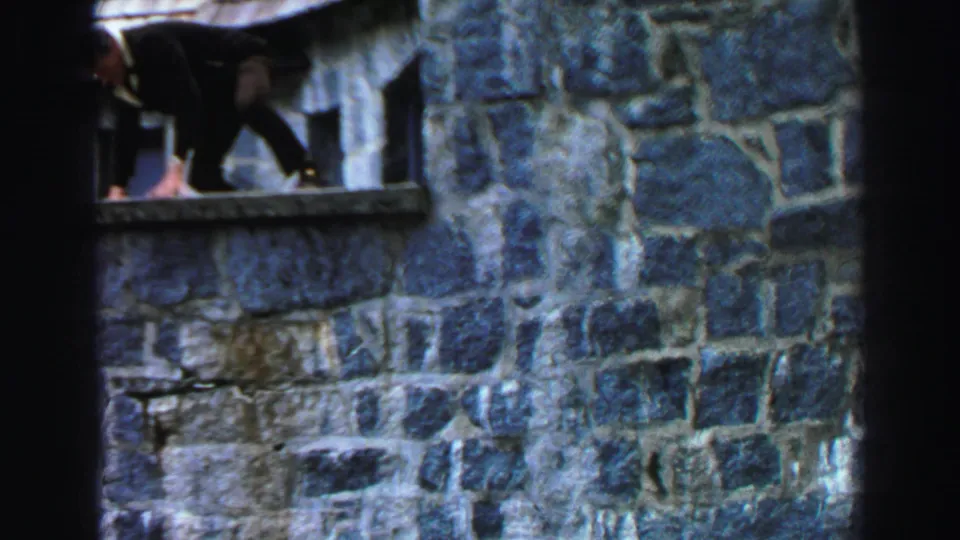 1969 Lake Tahoe home-movie frame showing the suited man climbing through a stone window opening in a rustic wall.