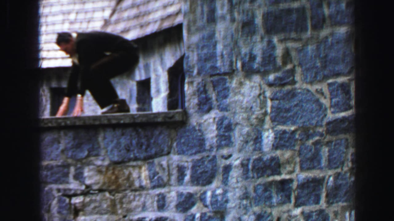 Man in dark clothing crouches on a stone cabin ledge beneath a shingled roof.