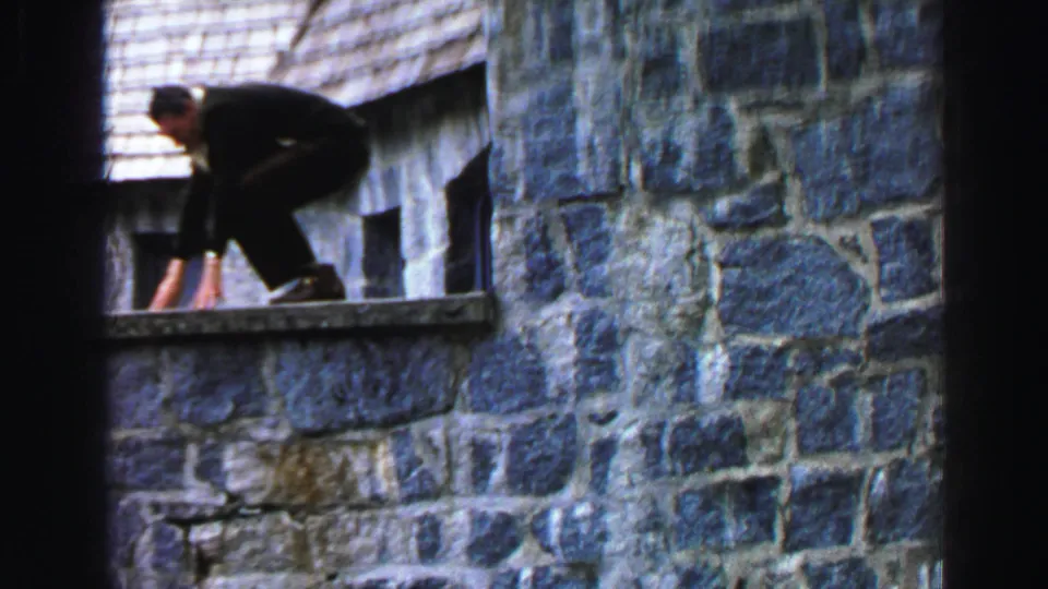 In 1969 Lake Tahoe home-movie footage, a man in dark clothing crouches on a narrow ledge along a blue-gray stone cabin wall.