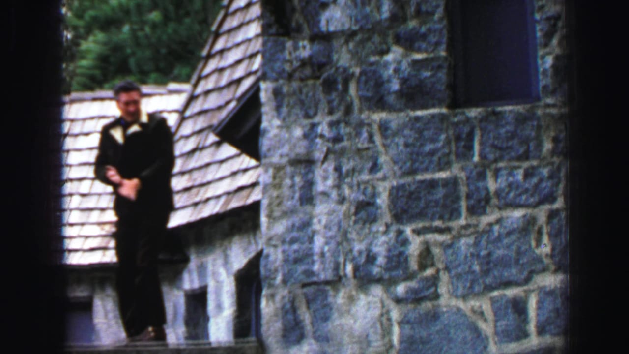 Man stands on a narrow ledge beside a stone cabin wall and shingled roof.