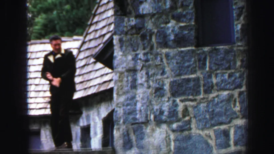 A suited man stands on a narrow exterior ledge of a Lake Tahoe stone cabin, with shingled rooflines and trees behind him.