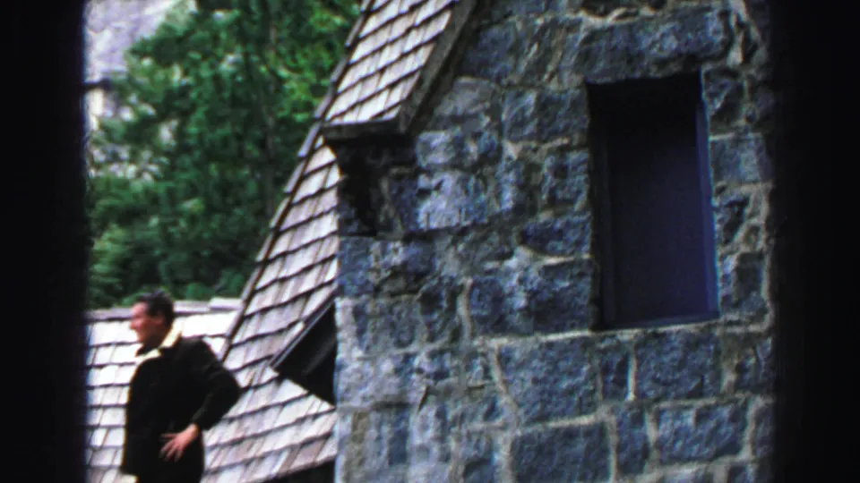 The man continues along the Lake Tahoe cabin exterior, partly framed by dark film borders, with stonework, roof shingles, and trees visible.