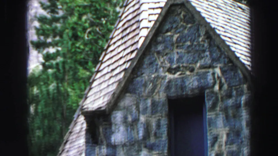 A Lake Tahoe stone cabin gable fills the frame, showing blue-gray masonry, pale roof shingles, a dark window opening, and forest behind.
