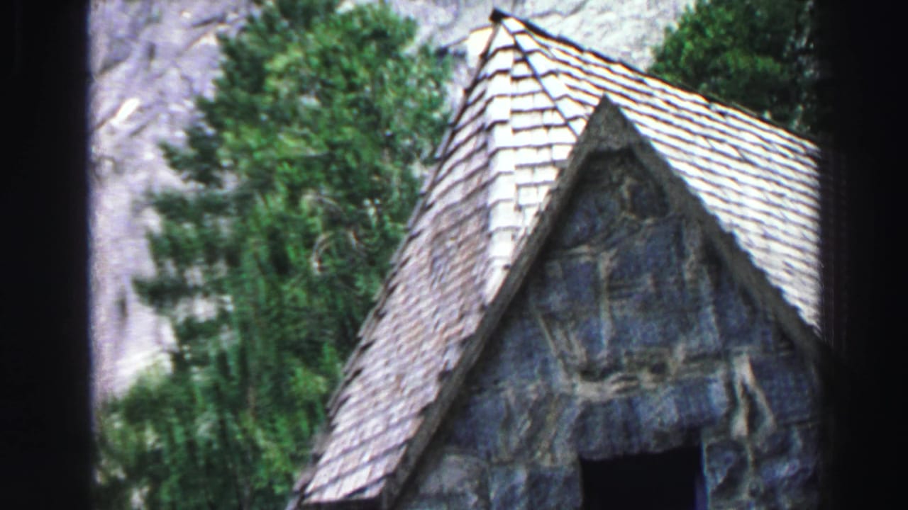 Stone gable building with a shingled roof among trees below granite cliffs.