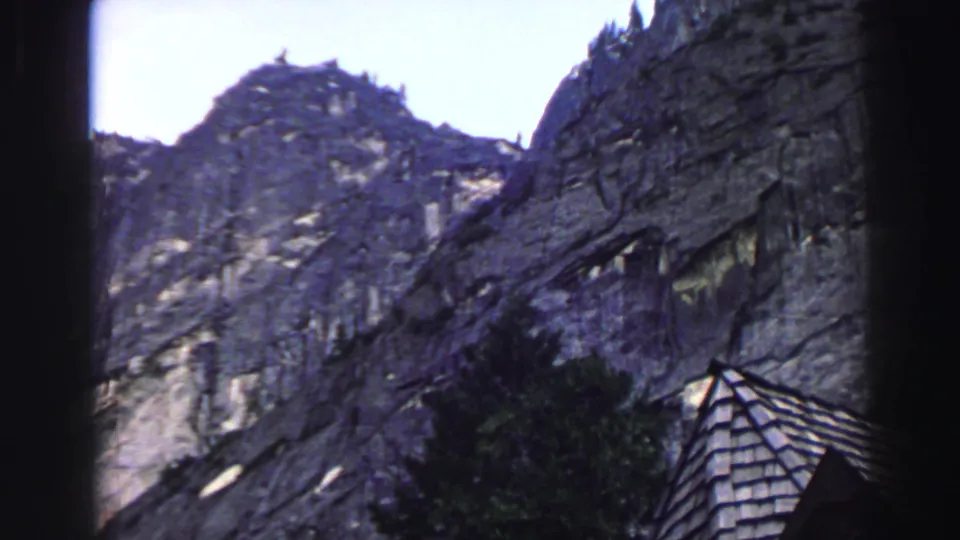 1969 Lake Tahoe 8mm frame of rugged granite slopes, evergreens, and the lower edge of a rustic shingled roof.