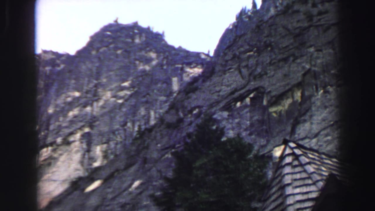 Steep rocky mountainside with dark trees and a small shingled roof at the lower edge.