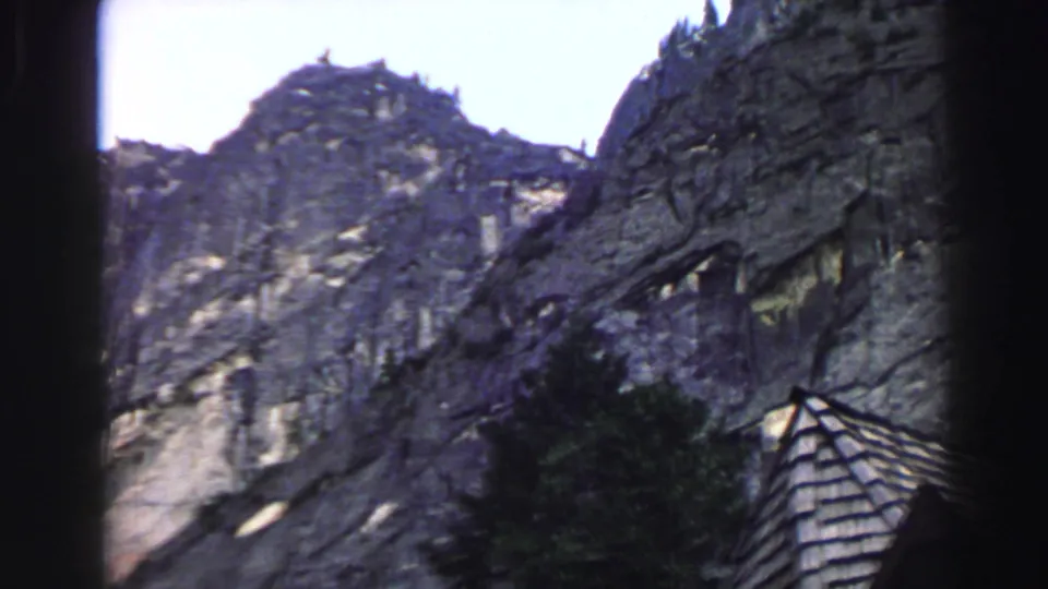1969 Lake Tahoe 8mm frame showing a steep gray rock face, dark pine foliage, bright sky, and a small shingled roof near the bottom.