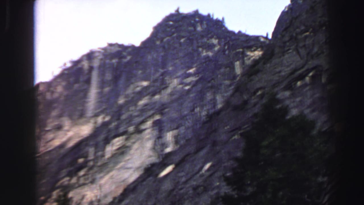 Rocky mountainside under bright sky with dark foliage along the lower right edge.