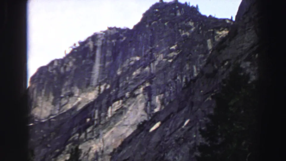 1969 Lake Tahoe 8mm frame showing a tall gray mountainside, dark evergreen trees, bright sky, and small pale patches on the slope.