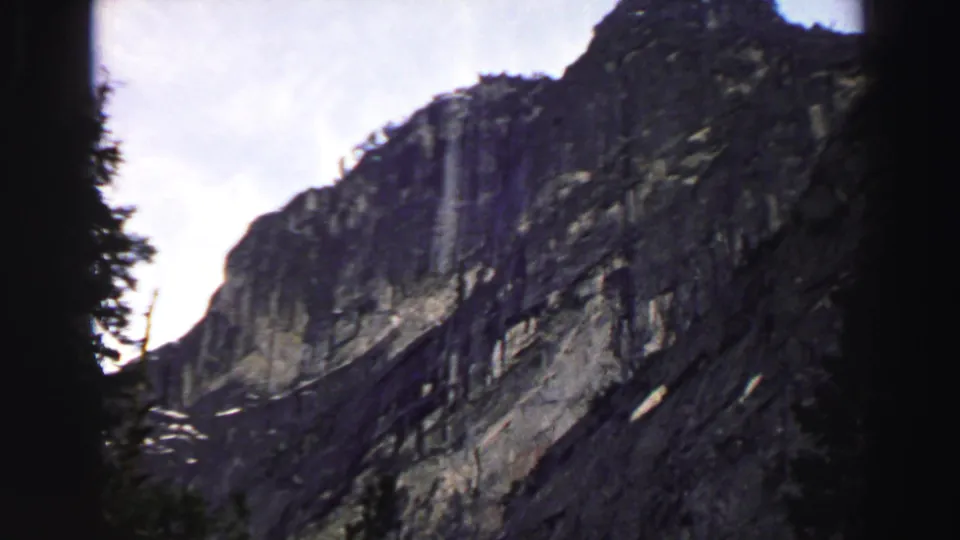 1969 Lake Tahoe 8mm frame with a rocky mountain slope, dark pine silhouettes, pale rock patches, and bright sky framed by film edges.