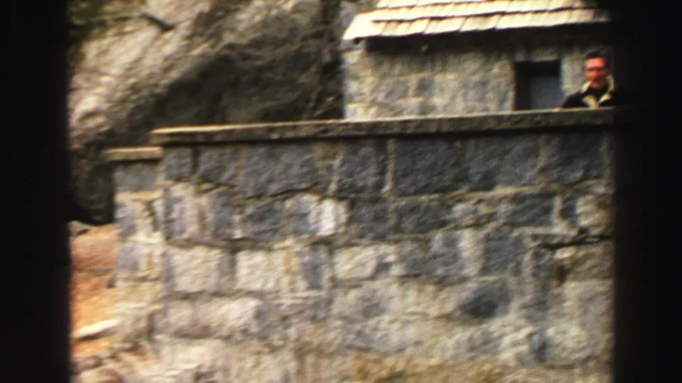 1969 Lake Tahoe home-movie frame showing a suited man behind a high stone wall beside a rustic building with a shingled roof.