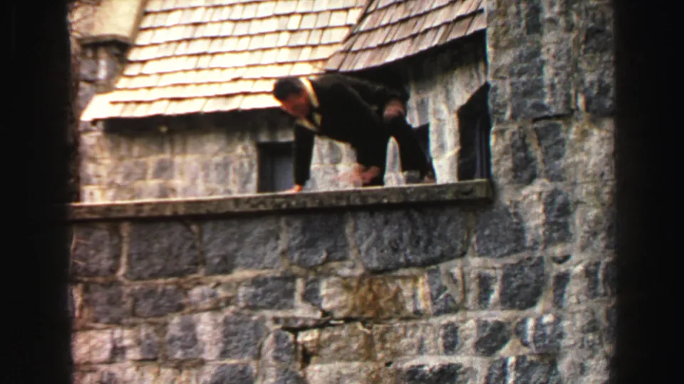 1969 Lake Tahoe footage shows the suited man crouched on a high stone wall, balancing beside the rustic building exterior.
