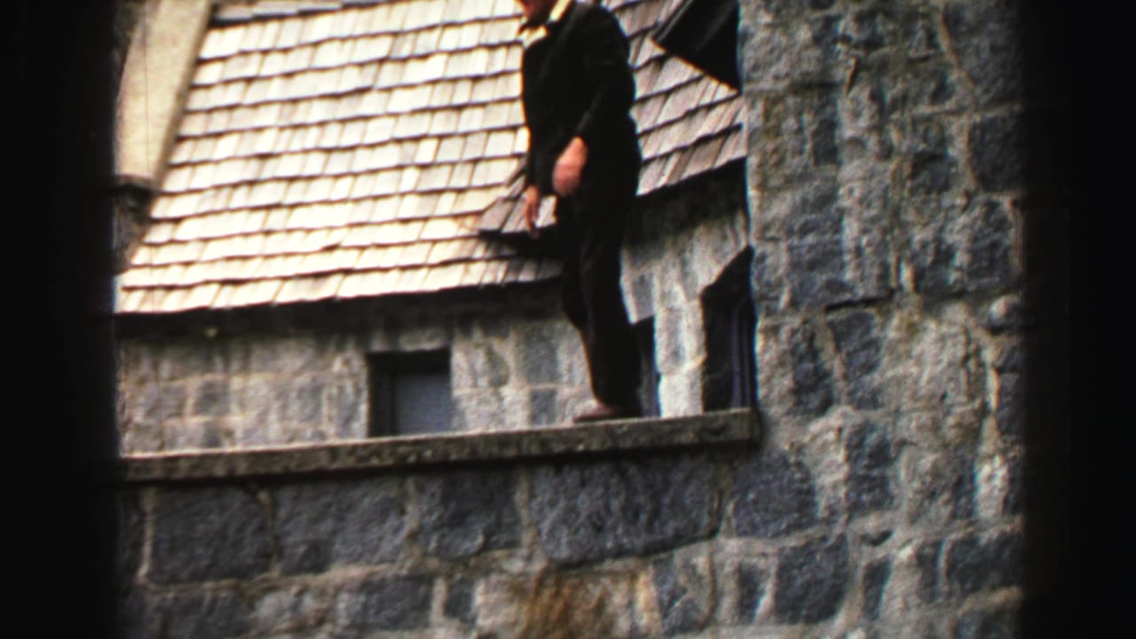 Man in a dark suit standing on a narrow stone ledge beside a shingled roof.